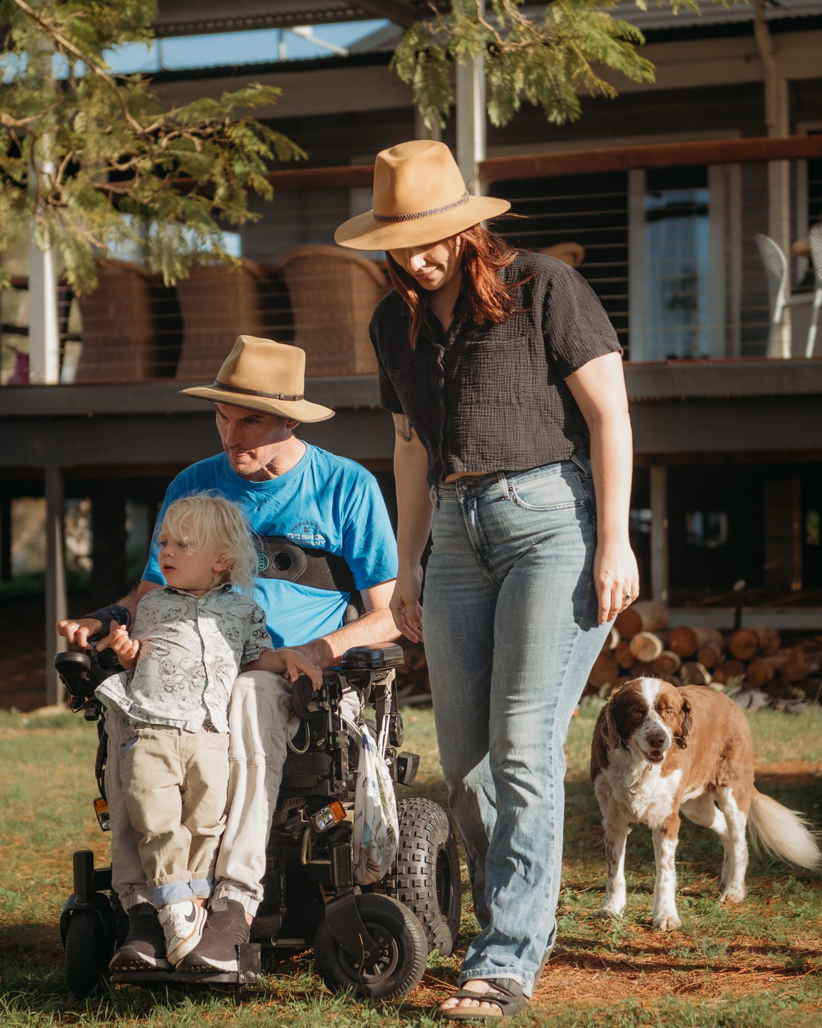 a man in an electric wheelchair with his son on his feet and his family and his dogs walking under a jacaranda tree